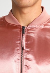 Rosa Satin-Bomberjacke mit durchgehendem Reißverschluss, geripptem Kragen und glatter Textur. Verfügt über einen schwarzen Reißverschluss mit elegantem Design.