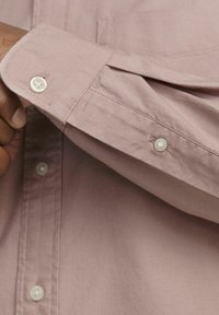 Camisa de manga larga rosa claro con puños abotonados. Presenta una tela suave y texturizada y un sutil detalle de bolsillo en el pecho.