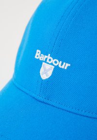 Boné azul feito de tecido texturizado, com o logótipo "Barbour" bordado em branco e detalhe de escudo na frente. Inclui aba curva.