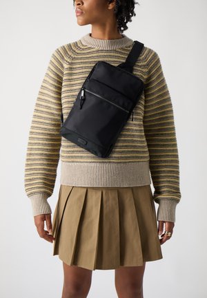 Sac bandoulière noir en nylon, de forme rectangulaire, avec deux poches zippées à l'avant. Le modèle porte un pull en maille rayé et une jupe plissée beige.