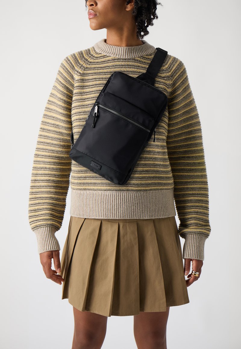 Sort nylon netpose, rektangulær form, med to forreste lynlåslommer. Modellen har en stribet striktrøje og en plisseret beige nederdel på.
