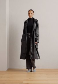 Svart trench coat i konstläder med avslappnad passform, stora kragar och dubbelknappning. Bärs över en svart polotröja och grå jeans.