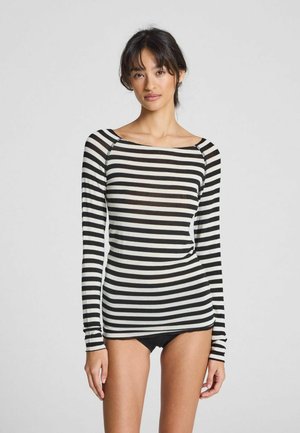 Vrouw in een zwart-wit gestreepte lange mouwen top en zwarte onderkleding, staande tegen een effen lichte achtergrond.