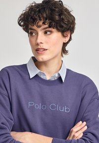 Lila sweatshirt med texten "Polo Club" i ljusblått, bärs över en blå och vit randig skjorta med krage. Slät textur, avslappnad design.