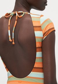 Maillot de bain rayé dans des tons orange, bleu et marron ; présente un dos nu avec un lien à nouer et des manches courtes. Tissu texturé avec des rayures horizontales.