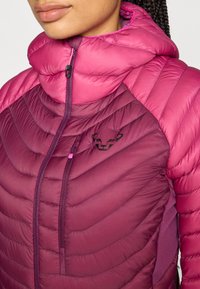 Chaqueta aislante en fucsia y burdeos con un alto cuello, costura en forma de espiga y un logo negro en el pecho. Bolsillo con cremallera en el lateral.