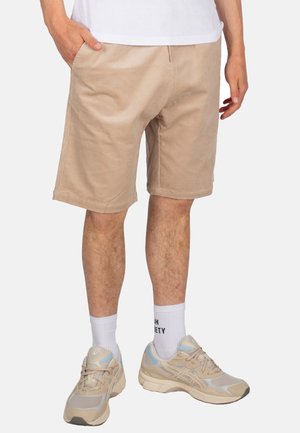 Shorts in cotone a coste beige con vestibilità relaxed, caratterizzati da tasche laterali e una texture liscia, abbinati a scarpe sportive beige e calzini bianchi alla caviglia.