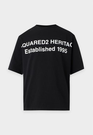 Jeune homme portant un t-shirt noir avec un texte blanc et un jean bleu délavé avec des détails de patchs, debout devant un fond clair uni.