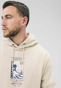 Mand med kort mørkt hår og skæg iført en beige hættetrøje med en blå og hvid bølgedesign samt japansk tekst på fronten.
