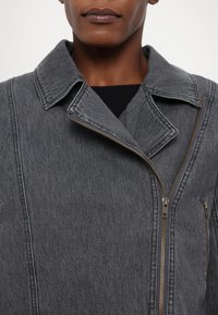 Giacca in denim grigio con colletto a punta, chiusura asimmetrica con zip e due tasche laterali, caratterizzata da una superficie leggermente strutturata.