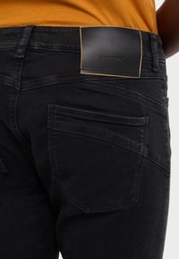 Black denim jeans feature a smooth texture, five-pocket design, and a black leather patch at the waist with embossed branding.