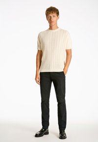 Maglione a maniche corte in crema con lavorazione a trecce e collo rotondo. Indossato con pantaloni neri aderenti e scarpe nere lucide.