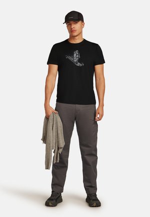 Zwarte katoenen t-shirt met een witte afdruk van een figuur die een golf berijdt. Gecombineerd met grijze cargobroek en een grijze sweater in de hand. Zwarte pet en schoenen.