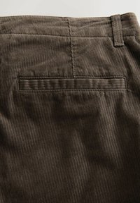 Dunkelbrauner Cordstoff mit einer horizontalen Paspeltasche und einem Gürtelschlaufen am oberen rechten Eck.