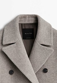 Manteau en laine gris avec de larges revers, deux boutons noirs et une étiquette intérieure noire indiquant Massimo Dutti fabriqué au Portugal.