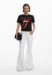Black cotton t-shirt with red Rolling Stones logo, paired with white flared jeans. Model holds a black bag. Simple background.