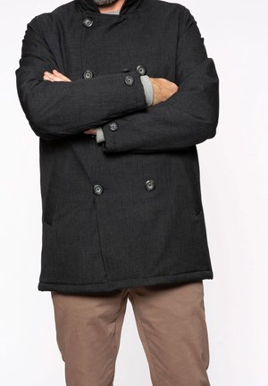 Person mit verschränkten Armen, trägt einen dunklen, zweireihigen Mantel und hellbraune Hosen vor einem schlichten Hintergrund.