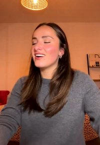 Jonge vrouw met lang bruin haar in een grijze trui glimlacht met gesloten ogen binnenshuis onder warm licht.