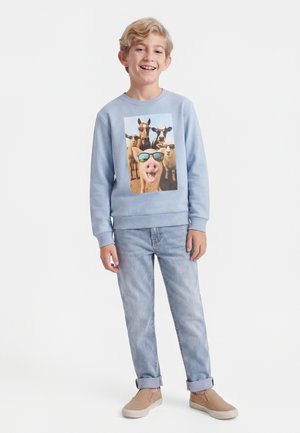 Smilende dreng iført en lyseblå sweatshirt med et tryk af en gris og gårddyr med solbriller, lyse jeans og beige slip-on sko.