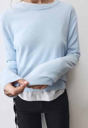 Personne ajustant la manche d'un pull bleu clair par-dessus une chemise blanche, portant un pantalon noir, debout devant un fond blanc uni.