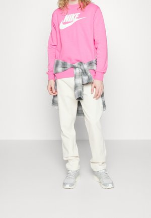 Person iført en lyserød Nike sweatshirt, hvide bukser, grå ternet skjorte bundet om livet og grå sneakers på en ensfarvet baggrund.