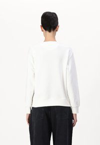 Personne aux cheveux courts et foncés portant un sweat-shirt blanc uni à manches longues et un pantalon noir, vue de dos sur fond blanc.