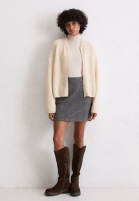 Cardigan lavorato a maglia crema, gonna grigia con zip, stivali al ginocchio in suede marrone. Texture morbide, vestibilità rilassata e palette di colori neutri.