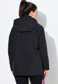 Sort vandtæt jakke med hætte. Har en glat tekstur, lange ærmer og en lidt løs pasform, der viser et minimalistisk design.
