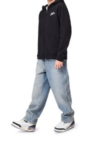 Person in einem schwarzen Zip-Hoodie, hellblauen weiten Jeans und weißen Turnschuhen, der mit einem Fuß leicht nach vorn steht.