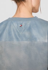 Top de tela perforada azul claro que presenta una sección superior lisa y ligeramente brillante, con un pequeño logotipo de estrella en la parte posterior del escote.