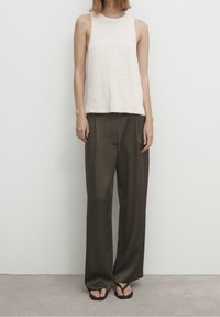 Débardeur en maille beige clair avec un col rond, porté avec un pantalon ample vert olive et des tongs noires, posé sur une surface neutre.