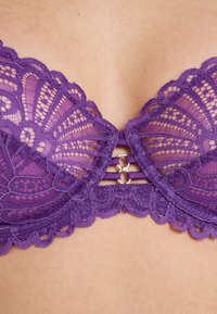 Reggiseno in pizzo viola con intricati motivi floreali, bordi scallop e una chiusura centrale in metallo. Tessuto liscio con una texture trasparente.