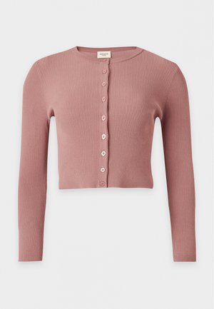 Femme portant un haut rose court à manches longues avec boutons, pantalon large bleu foncé brodé, et sandales plates marron, debout les mains dans les poches.