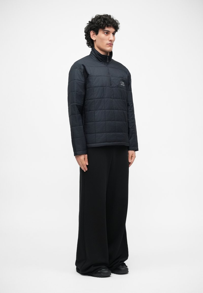 Black quilted pullover jacket with a high collar, zip closure, and front pocket. Worn with oversized black trousers and dark shoes.