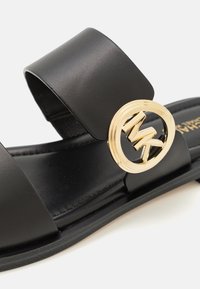 Sandale noire avec une large bride, texture lisse et accent logo doré circulaire. Semelle plate avec un design de semelle minimal.
