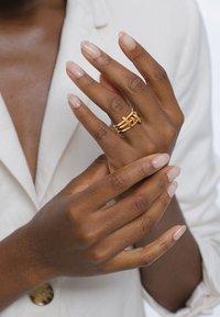 Goldene Multi-Band-Ringe mit verflochtenen Designs. Hände mit neutralem Nagellack, die vor einem hellen Stoffhintergrund positioniert sind.