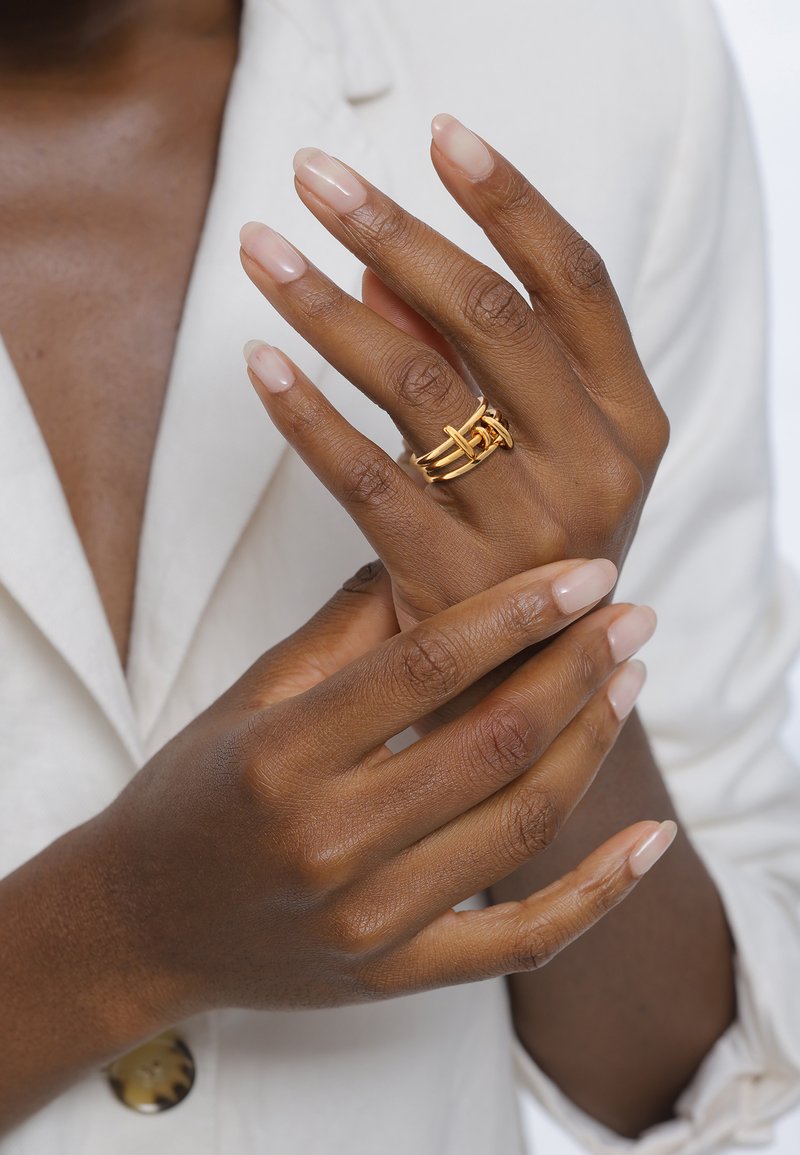 Goldene Multi-Band-Ringe mit verflochtenen Designs. Hände mit neutralem Nagellack, die vor einem hellen Stoffhintergrund positioniert sind.