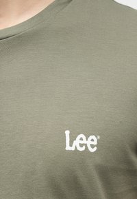 Κοντινό πλάνο υφάσματος σε ελιά πράσινο με το λευκό λογότυπο «Lee» τυπωμένο στην περιοχή του στήθους σε μπλούζα με στρογγυλή λαιμόκοψη.