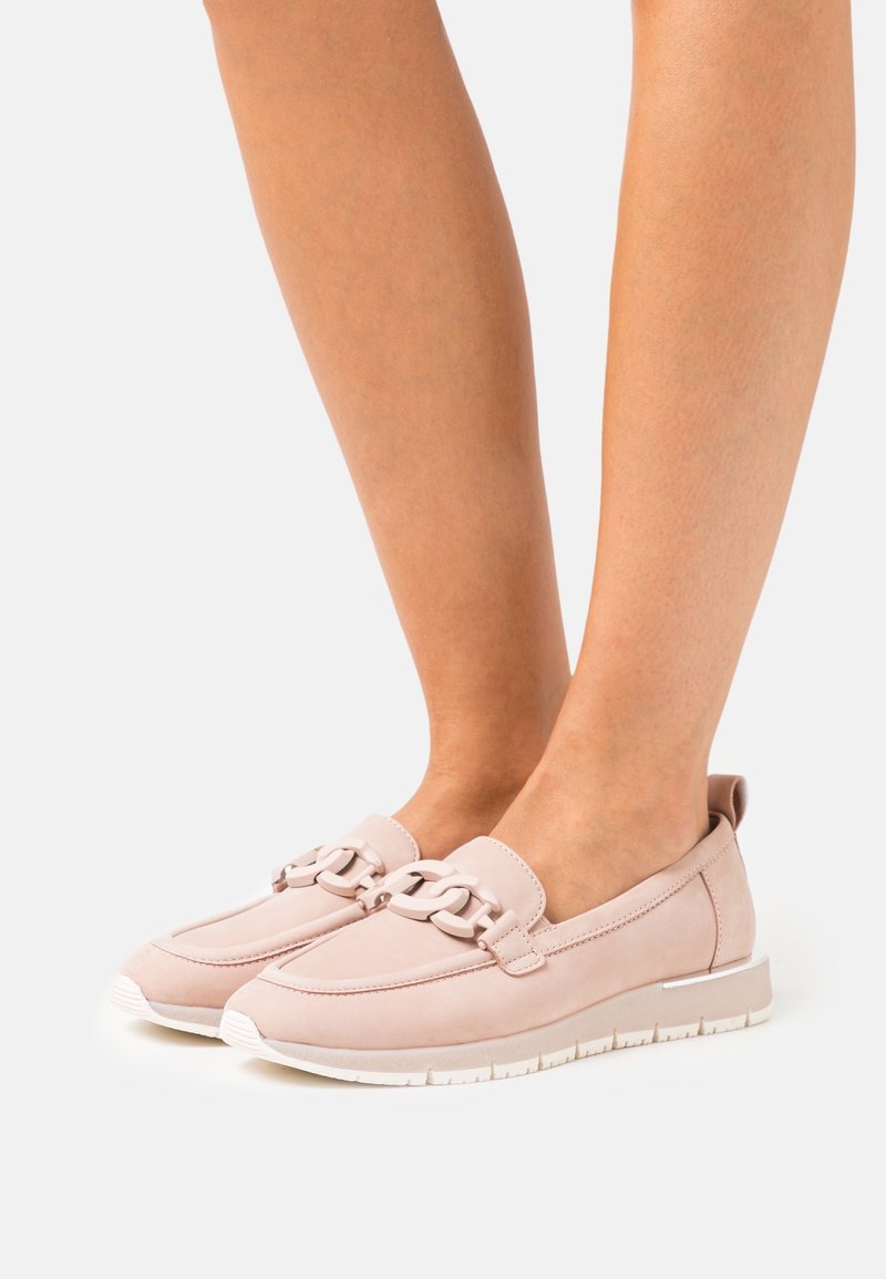 Jambes nues portant des mocassins légers rose clair à enfiler avec un gros détail chaîne sur le dessus et des semelles blanches en caoutchouc sur fond blanc.