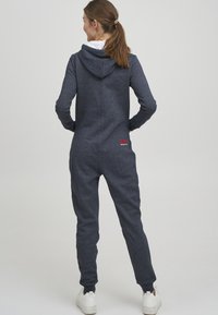 Mørkeblå hette-jumpsuit laget av myk, teksturert stoff. Har glidelås foran og elastiske mansjetter, med en liten logo bak.