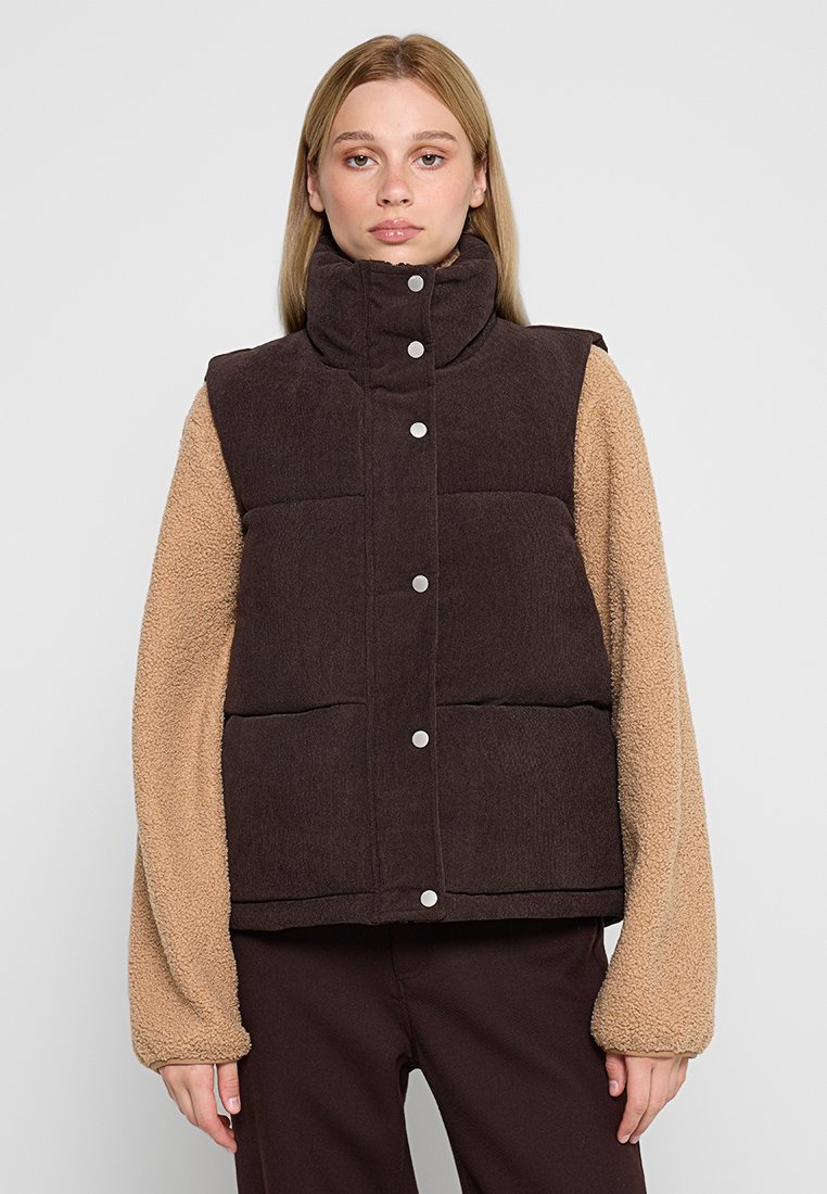 Braune gepolsterte Weste mit hohem Kragen und silbernen Druckknöpfen. Ärmel aus tanfarbigem Plüschstoff. Texturiertes Material und zweifarbiger Design.