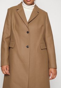 Brown wool-blend coat with a tailored design, notched lapels, two button closures, and side pockets. Smooth texture with subtle herringbone pattern.