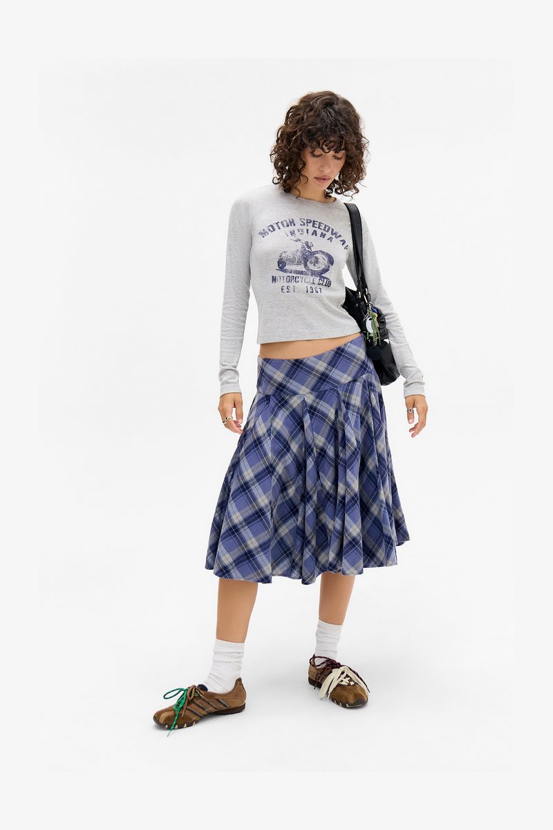 Camisola cinza de manga longa e cortada com gráfico de motocicleta azul; saia de calças rodada em padrão xadrez azul; sapatos castanhos com atacadores coloridos; meias brancas.