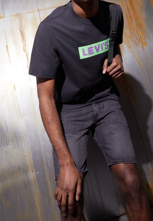 Homme portant un t-shirt noir Levi's et un short en denim gris foncé, ajustant la sangle d'un sac noir, debout devant un mur en métal rouillé.