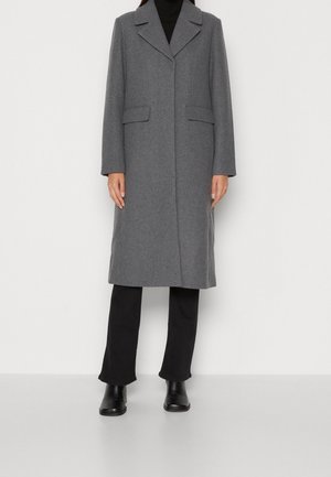 Kvinde iført en lang grå uldfrakke over sorte bukser og sorte ankelstøvler, stående mod en ensfarvet hvid baggrund.