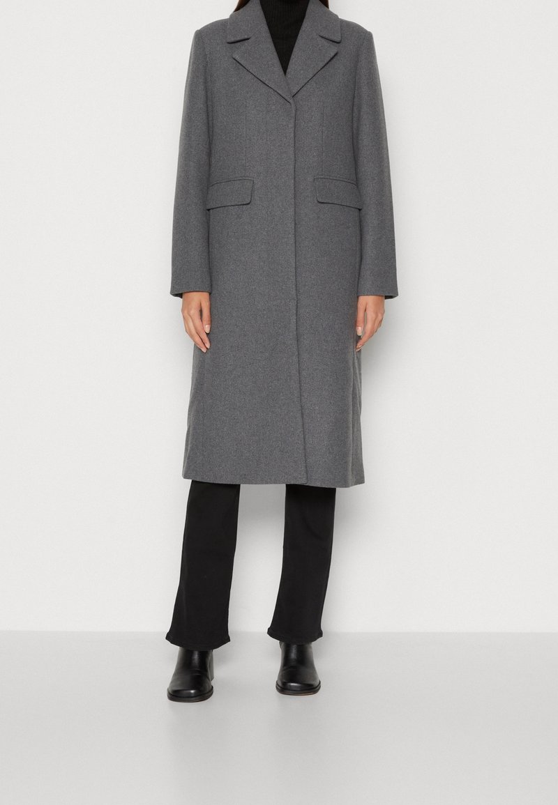 Femme portant un long manteau gris en laine par-dessus un pantalon noir et des bottines noires, debout devant un fond blanc uni.