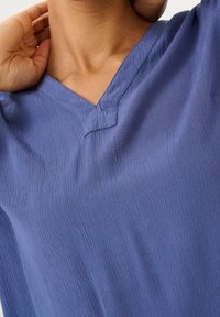 Haut bleu à col en V en tissu texturé, présentant un motif côtelé subtil et des manches courtes. L'encolure est finie par une couture nette.