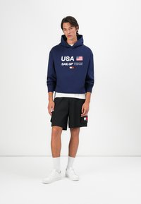 Felpa cropped blu navy con la scritta "USA SAIL GP TEAM", abbinata a pantaloni corti neri con una toppa. Sneakers bianche e calze bianche a costine completano il look.
