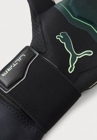 Svart målvaktshandske med texturerad tyg, gröna detaljer och en framträdande vit Puma-logotyp på baksidan.