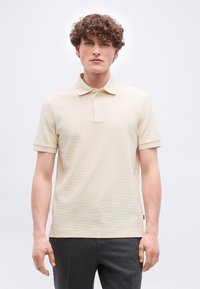 Νεαρός άνδρας με σγουρά μαλλιά, φορώντας μπεζ κοντομάνικο πόλο με υφή και σκούρο γκρι παντελόνι, στέκεται μπροστά σε μονοχρωμο φόντο.